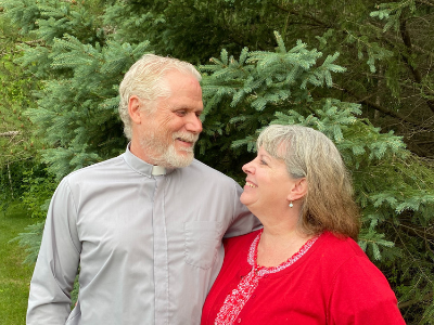 catholic deacon joe utecht smiles at his wife, margaret. there are green pine trees in the background